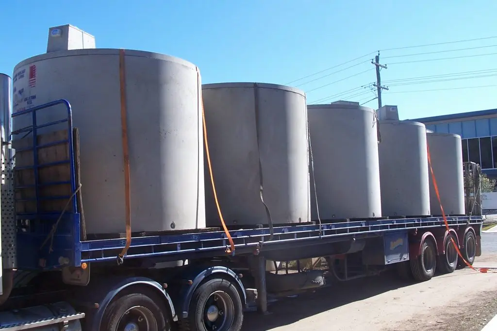 A flatbed truck carrying several large cylindrical septic tanks secured with orange straps. The tanks are lined up horizontally on the truck bed. In the background, there are industrial buildings and power lines against a clear blue sky.