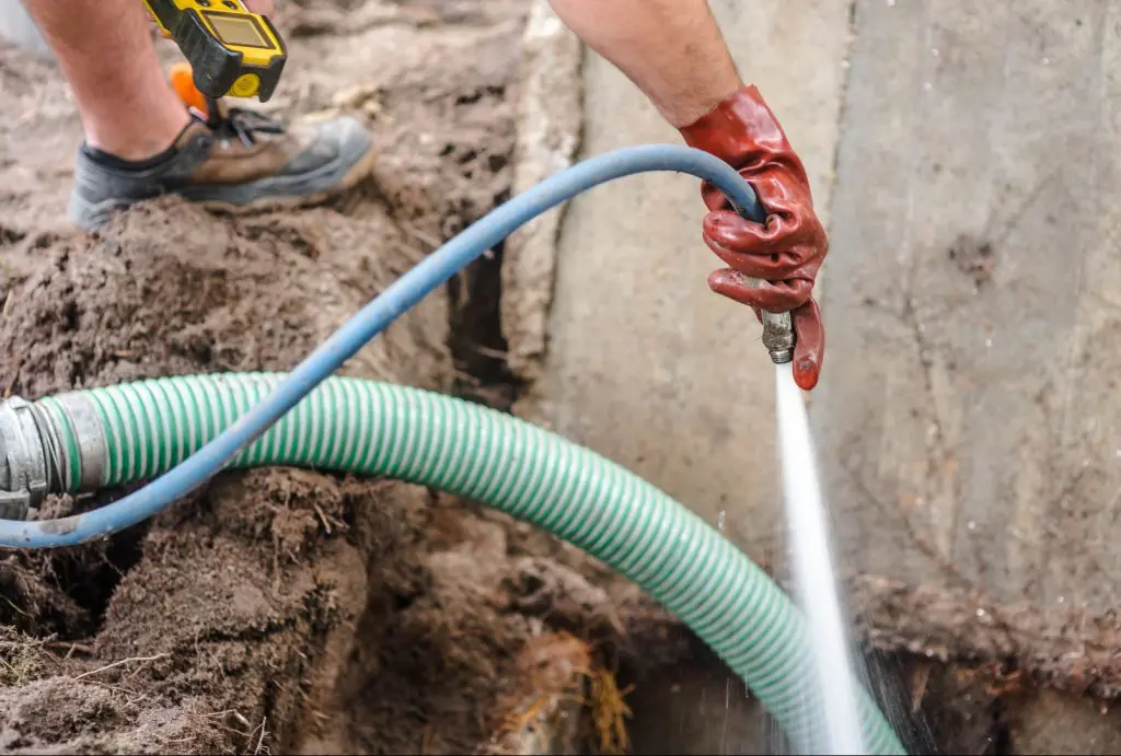 A person wearing red gloves and work boots uses a blue hose to spray water near a green striped hose in a partially muddy area. The individual holds a device attached to their belt, likely for monitoring septic tanks. A concrete structure is visible in the background.