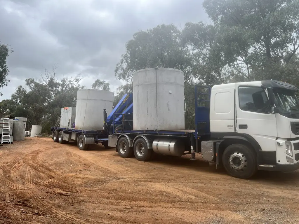 A truck with a trailer is transporting two large, cylindrical septic tanks on a dirt road, surrounded by trees and cloudy skies. The heavy-duty vehicle is equipped with lifting equipment for handling these essential components for domestic wastewater systems.