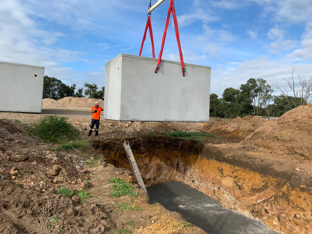 A construction worker, wearing an orange high-visibility jacket and helmet, observes as a large precast concrete structure for a domestic wastewater system is lifted by a crane over a prepared excavation site surrounded by dirt and greenery under a partly cloudy sky.