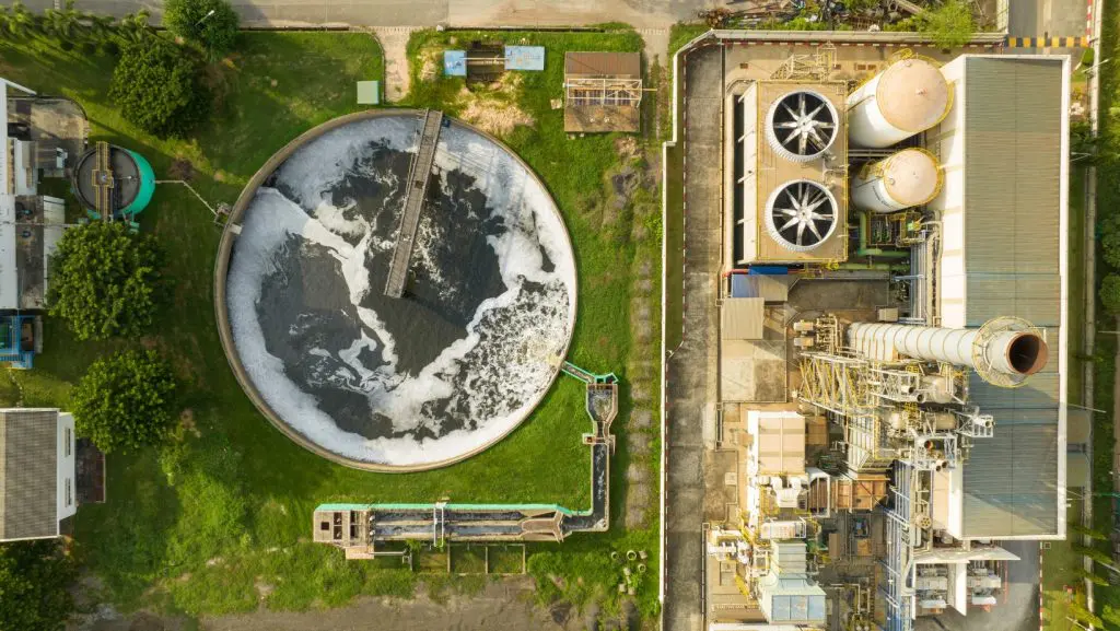 Aerial view of an industrial facility featuring a large circular water treatment tank with frothy water, integral to advanced wastewater systems. The scene includes green grass, pathways, and multiple structures with pipes and machinery, surrounded by various commercial buildings and equipment.