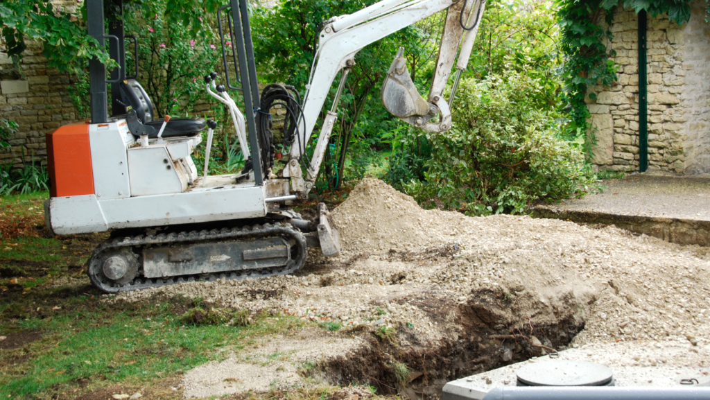 A small excavator with a white and orange body is digging a trench in a garden area surrounded by green plants and a stone wall. The excavator's bucket is filled with soil, creating a pile of dirt next to it. The scene suggests construction or landscaping work, possibly for installing septic tanks or wastewater systems.