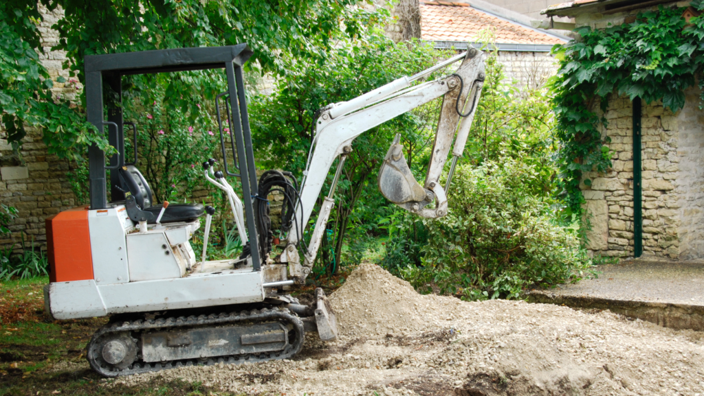 A small white and orange excavator is parked on a dirt patch in a garden area, surrounded by green trees and bushes. The excavator's arm is extended and its bucket is slightly raised. A stone wall and a building with a tiled roof are visible in the background, likely part of domestic wastewater systems maintenance.