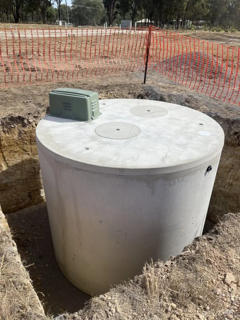 Image of a large cylindrical concrete tank partially buried in the ground at a construction site. The tank, part of a commercial wastewater system, has two access covers on top and a green electrical box attached to it. The area is surrounded by orange safety fencing and dry earth.