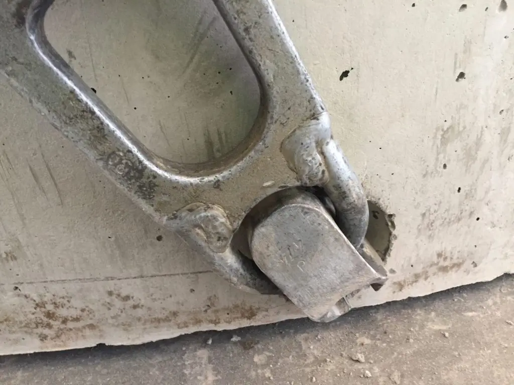 A close-up view of a heavy-duty metal shackle secured to a concrete surface, reminiscent of those found in wastewater systems. The shackle is weathered and shows signs of wear, indicating that it has been in use for a considerable time. The concrete around the shackle is also worn, with some small cracks and marks.