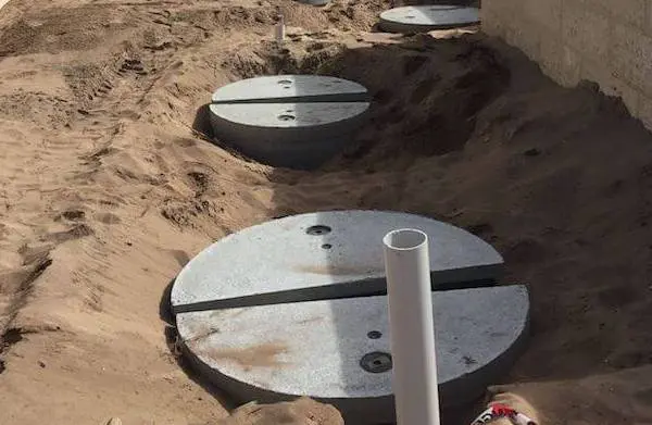 Circular concrete covers partially buried in sand, aligned vertically next to a concrete wall. PVC pipes are emerging from the center of each cover, indicating underground utility or wastewater systems. The area surrounding is slightly dug up, exposing more sand.