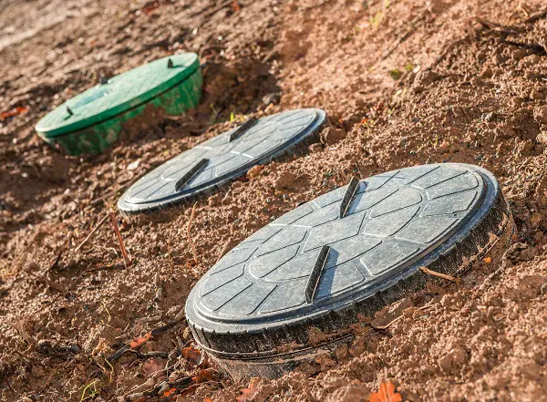 Three circular septic tank lids partially buried in the ground. The lids, essential to wastewater systems, rest on rough, dry soil and are arranged in a diagonal line. The closest lid is dark gray, the middle one is also dark gray, and the farthest lid is green.