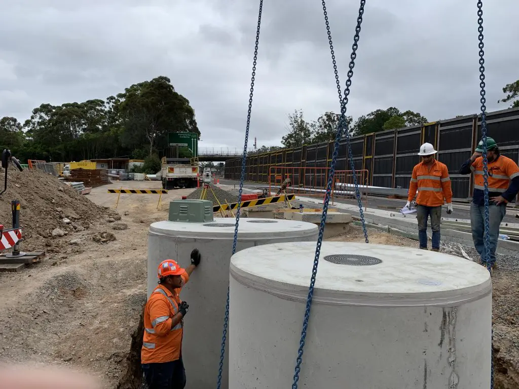 A construction site with three workers wearing orange safety vests and helmets. Two workers, one holding papers, stand on a raised platform while another operates a crane lifting large concrete septic tanks. Construction equipment and barriers are visible in the background.