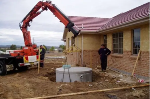 A crane mounted on a truck is installing a large round concrete septic tank into a hole next to a brick house. Two workers stand nearby, observing the process. The sky is overcast, and the ground is covered with scattered construction materials.