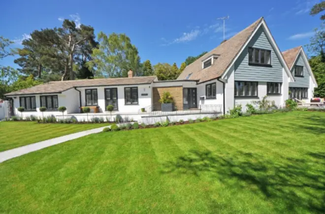 A large modern house with white walls and a green lawn under a bright blue sky. The home features multiple sections, varying heights, and numerous large windows. Surrounded by trees and greenery, a pathway leads to the main entrance, blending commercial elegance with domestic warmth.