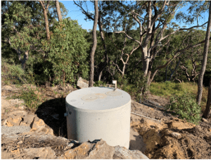 A circular, concrete water tank is partially buried in the ground, surrounded by trees and rocks in a forested area. The tank appears to be part of a domestic wastewater system, with some pipes and a valve on top. The scene is set in daylight with a clear blue sky.