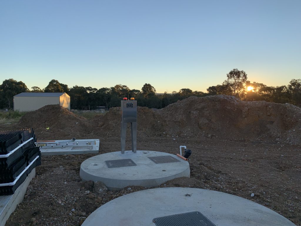 A construction site at sunset, featuring a concrete structure and metal equipment in the foreground. Piles of dirt and construction materials are visible, along with a small building and forested area in the background. The clear sky with the sun low on the horizon casts a warm glow over this commercial project.