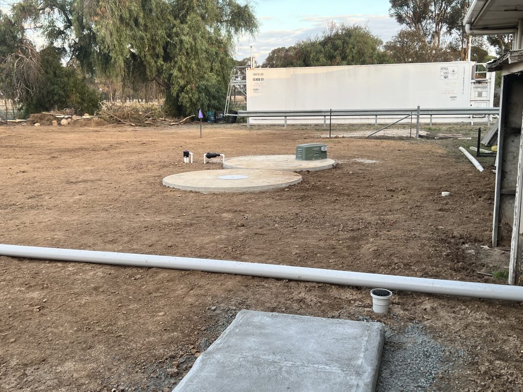 A construction site with an open dirt area showcasing two large, round concrete septic tank covers on the ground. Various pipes and a small green utility box are visible. In the background, there are trees and a large white structure, possibly a portable building.