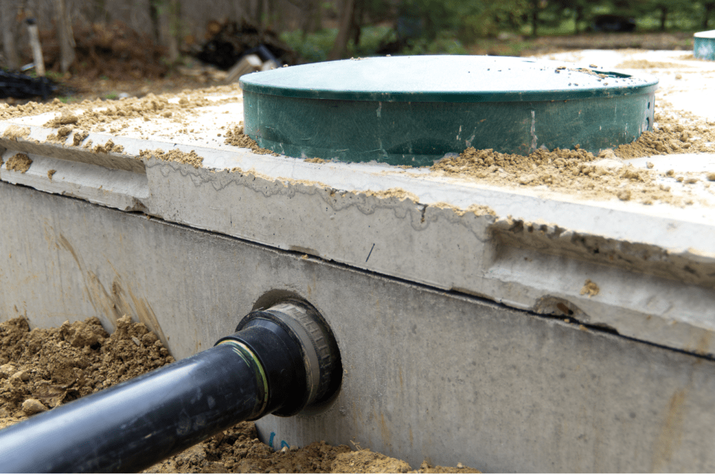 Close-up of a commercial concrete septic tank with a black pipe inserted into a sealed opening. The tank lid is visible at the top, partially covered with dirt. The surrounding area is filled with soil and outdoor vegetation, blurred in the background, showing part of a comprehensive wastewater system.