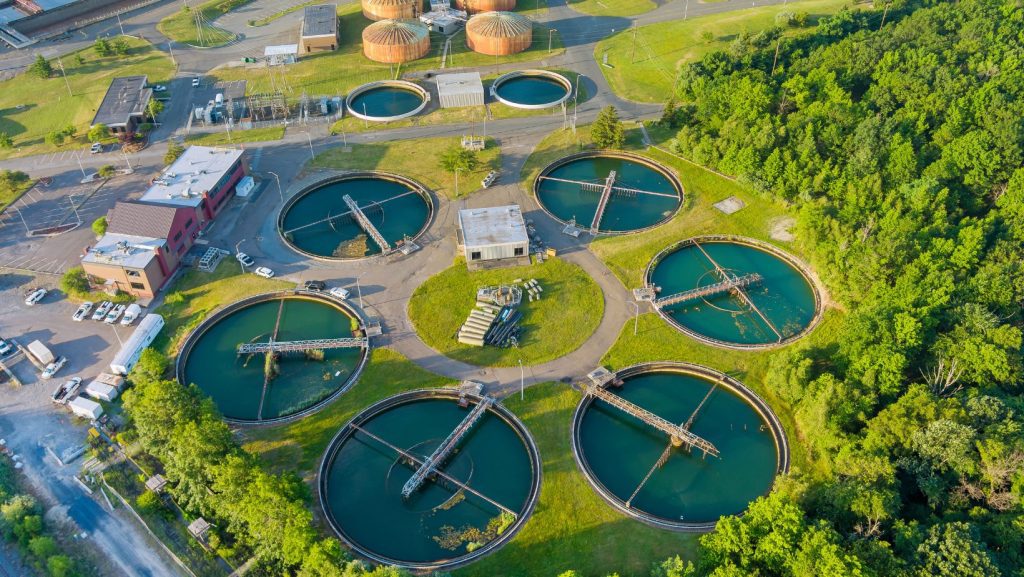 Aerial view of a wastewater treatment plant with six large circular settling tanks and various buildings. Four circular tanks occupy the central area, while two more are positioned above. This AWTS facility, surrounded by trees and greenery, features roads connecting the structures.