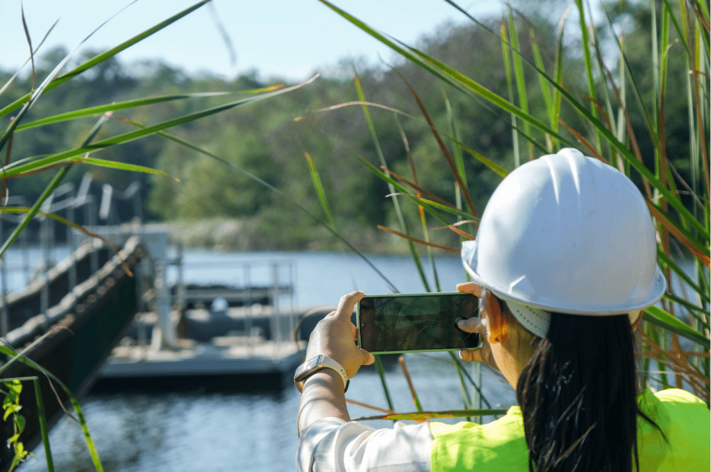 A person wearing a white hard hat and a yellow safety vest is taking a photo with a smartphone near a body of water. The background features an AWTS structure on the water and lush greenery. The person is partially obscured by tall grass in the foreground, highlighting the domestic utilization of wastewater systems.