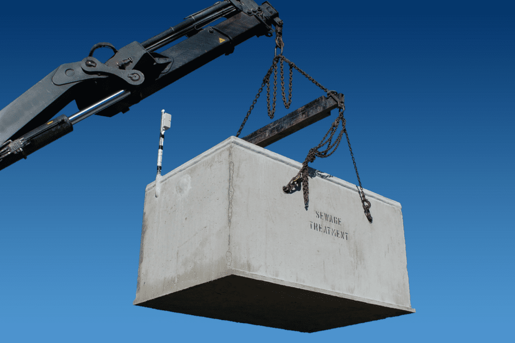 A large concrete block labeled "SEWAGE TREATMENT" is suspended in the air by chains attached to a crane against a clear blue sky background, showcasing the efficiency of modern wastewater systems.