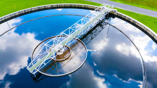 Aerial view of a wastewater treatment plant clarifier. The circular tank, resembling those used in commercial septic systems, has a white metal bridge-like structure across its diameter, with reflected clouds on the water surface. Green grass and a road surround the tank.