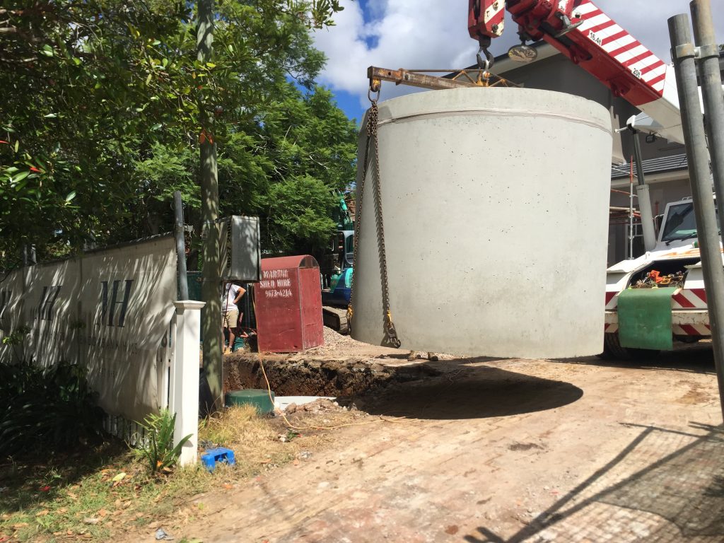 A large concrete septic tank, crucial for domestic wastewater systems, is being lifted by a crane onto a construction site. The area is enclosed by fencing, with construction equipment and a parked vehicle visible. Trees and a building are in the background, and a sign indicates a construction site.