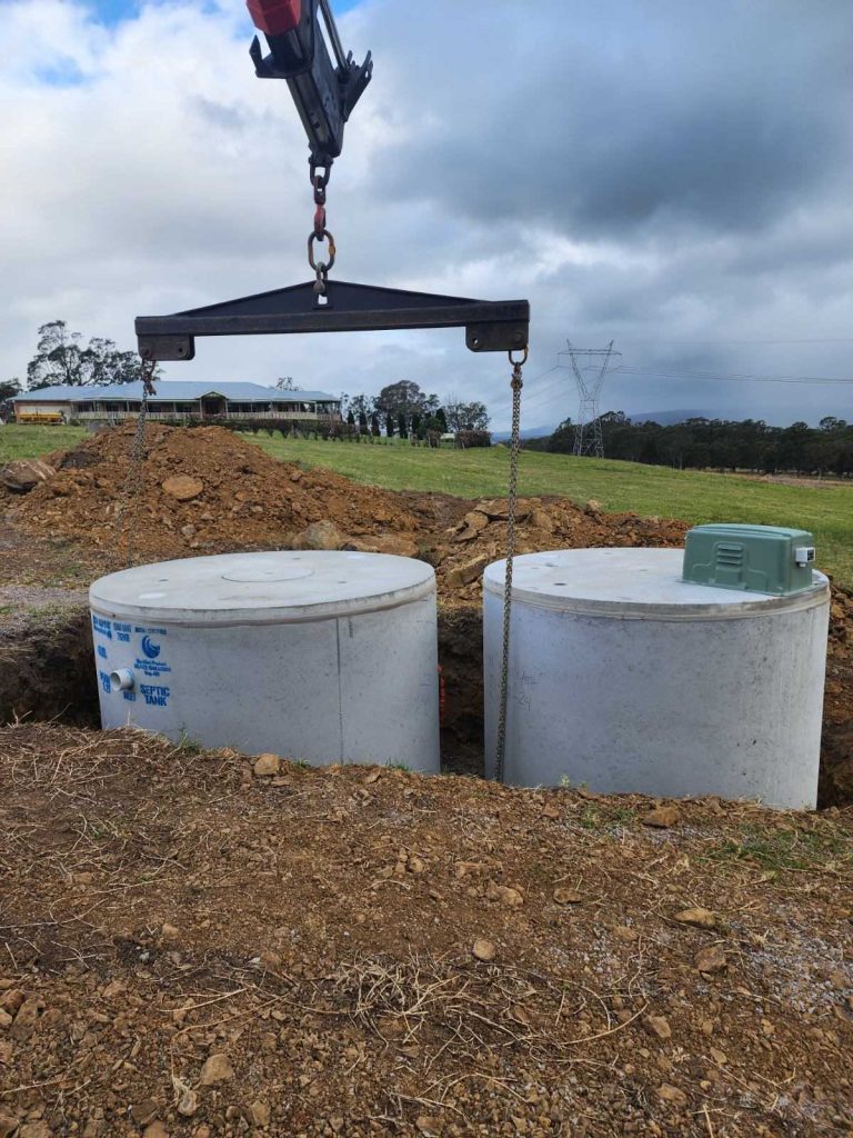 A crane is lowering two large cylindrical concrete tanks into a dug-up area at a construction site, likely for an AWTS designed to enhance wastewater systems. The surrounding area consists of dirt, grass, and some scattered rocks, with a few commercial buildings and power lines visible in the background under a cloudy sky.