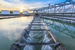 A water treatment facility with cascading water flowing over a series of metal barriers. The sunset reflects on the water, and a steel bridge structure spans the scene, with industrial tanks visible in the background.