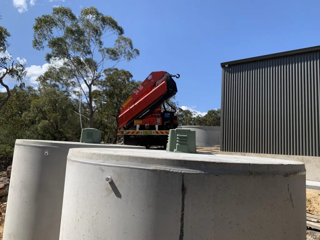 A construction site featuring a red crane truck positioned beside large concrete cylinders, part of a domestic wastewater system project, with trees and a building in the background under a clear blue sky.