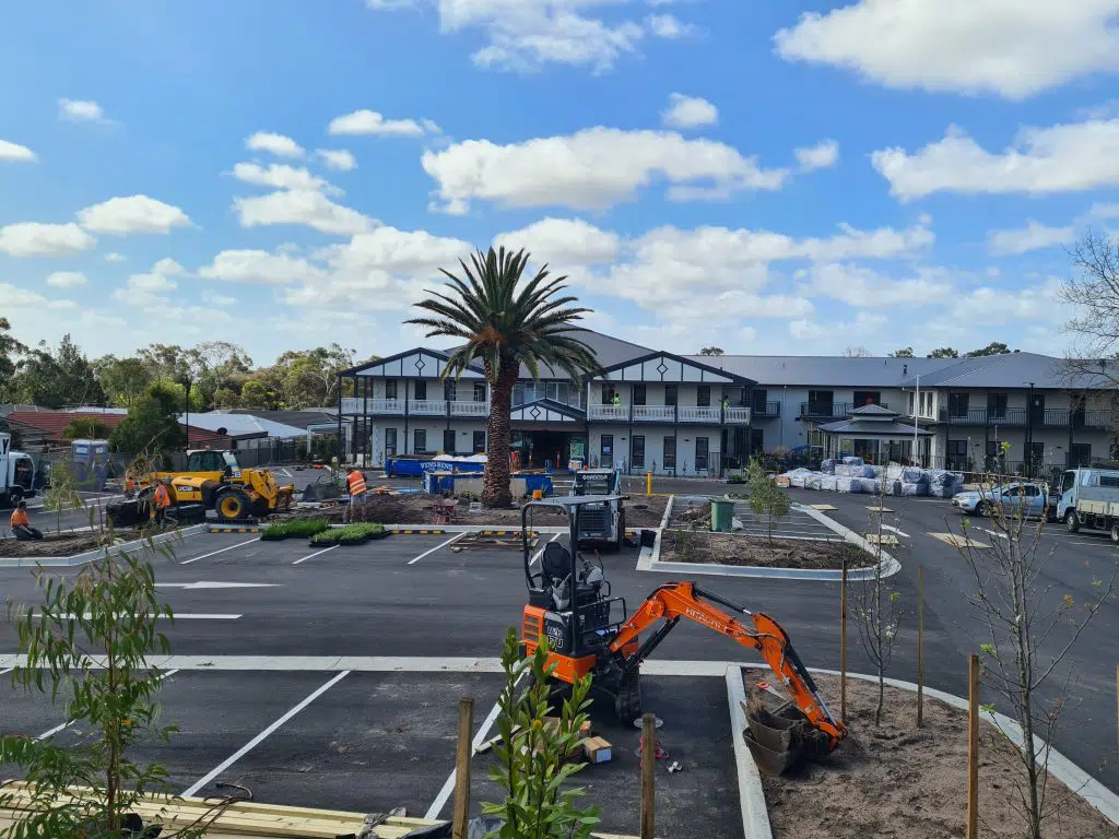 A construction site with several pieces of heavy machinery, including an orange excavator and a yellow backhoe loader, surrounded by an almost-completed parking lot. A large palm tree stands in the center. In the background are buildings with blue roofs, hinting at a nearby facility for domestic wastewater systems.