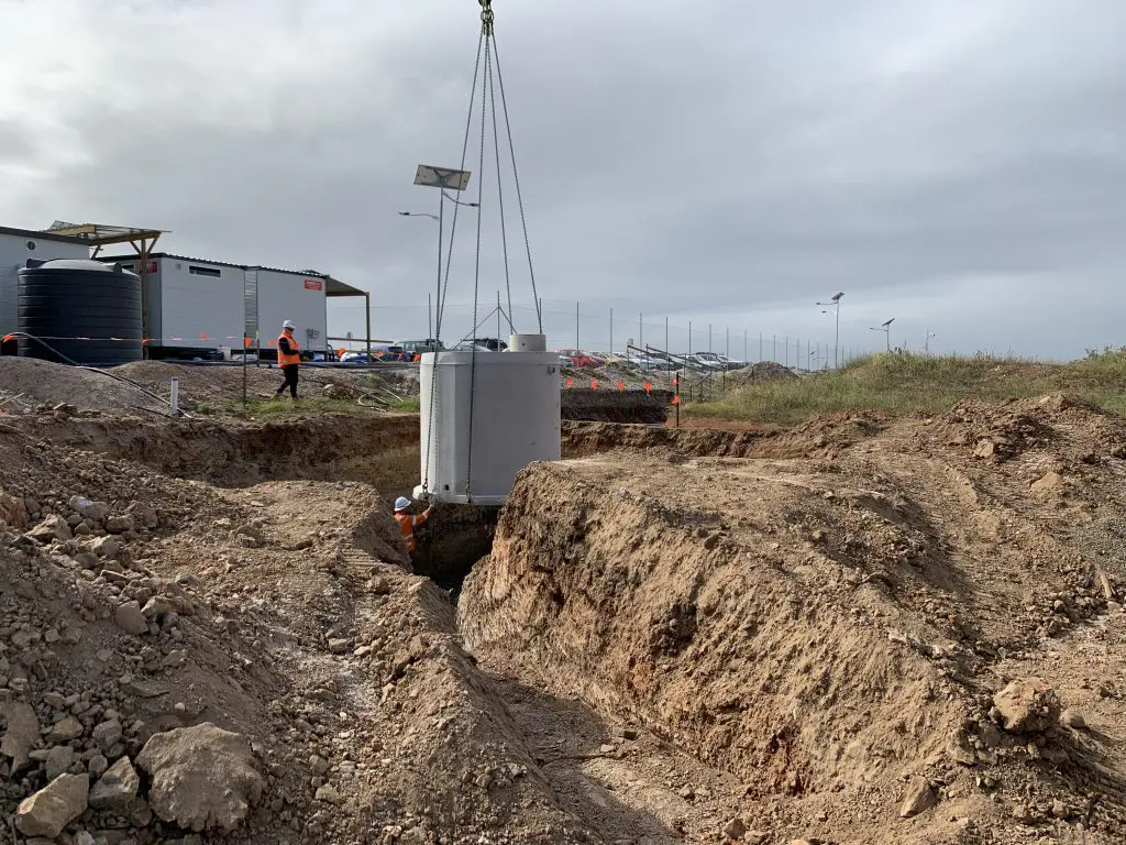 A large cylindrical concrete septic tank is being lowered into a trench by a crane at a commercial construction site. Visible are construction workers in safety gear, fencing, equipment, and a partially cloudy sky in the background. Dirt and soil surround the trench.