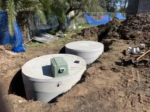 Two large concrete septic tanks, partially buried in a dirt area at a construction site, feature a green control box mounted on top of one. Nearby stands a palm tree, bordered by construction fencing and scattered debris. These domestic AWTS units are vital for efficient wastewater management.