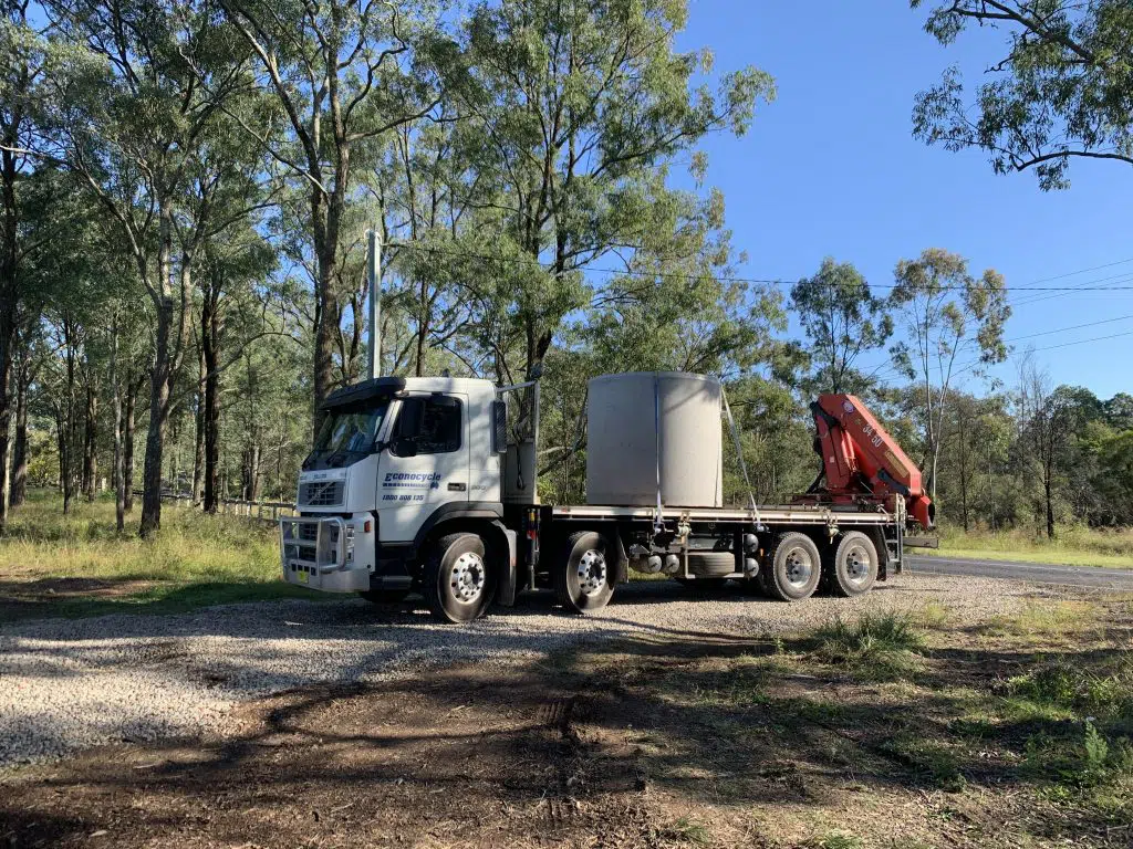 A large flatbed truck with a red crane arm is parked on a gravel road in a wooded area, transporting a large concrete pipe for wastewater systems. Trees and a clear blue sky are visible in the background.