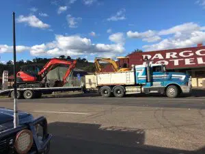 A blue and white truck is hauling two pieces of construction equipment, a red excavator and a yellow backhoe, on a flatbed trailer parked on a street in front of a building with a sign that reads "Dargo Hotel." The sky is partly cloudy, hinting at potential work involving septic tanks or wastewater systems.