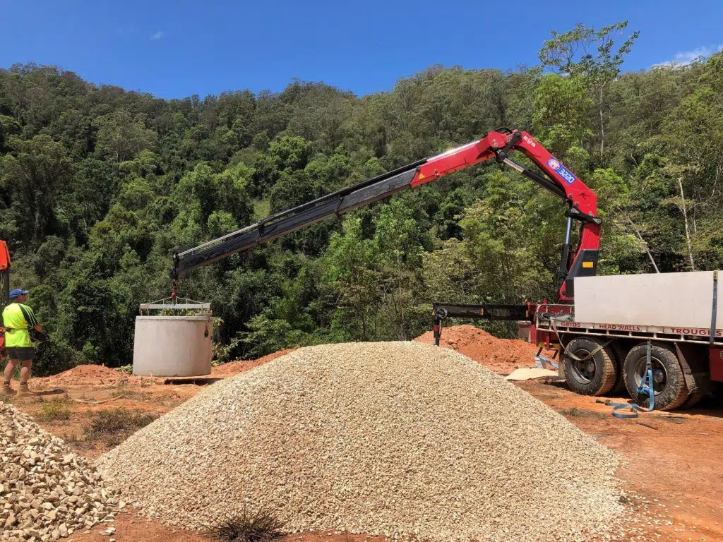 A construction site amidst a forested area. A worker in a yellow vest and hard hat stands near a large pile of gravel. A truck with a crane is lifting a large cylindrical concrete pipe for the domestic wastewater systems project. Trees and blue sky are visible in the background.