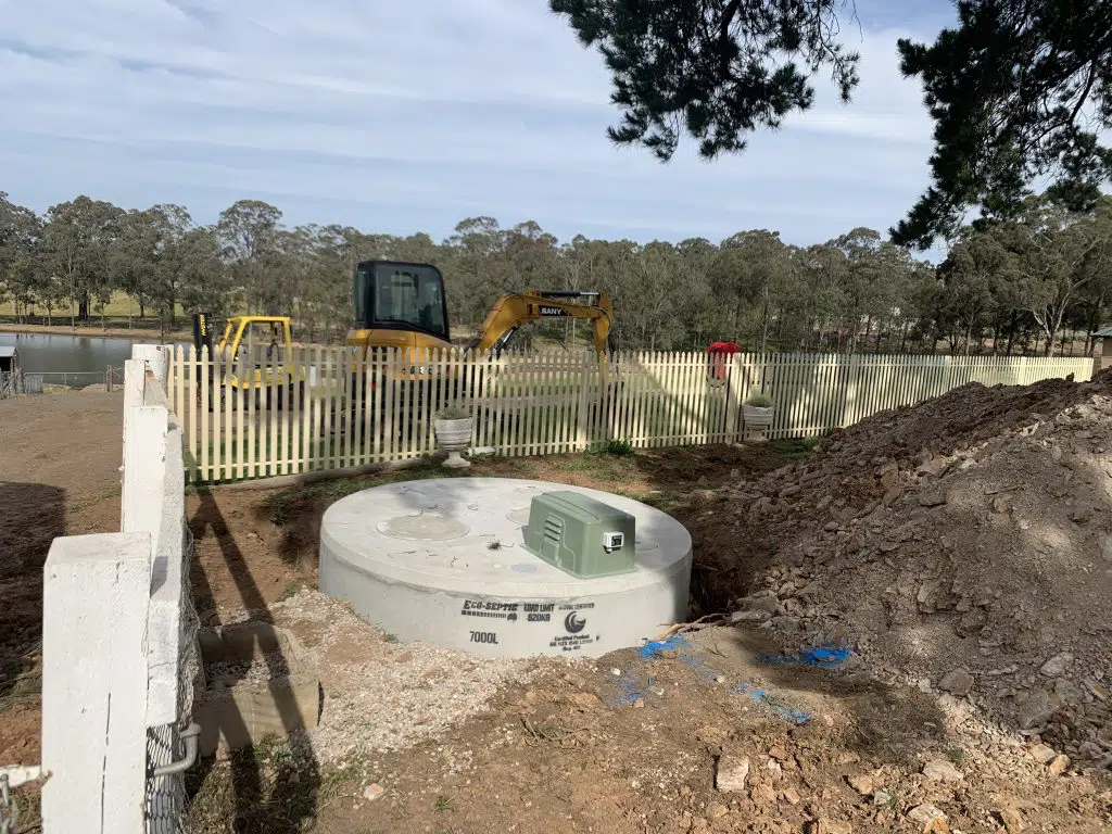 A construction site features a large, circular concrete septic tank partially buried in the ground, a green utility box on top, and a pile of dirt to the right. In the background, there is a yellow excavator next to a white picket fence and a scenic view of trees and a pond.