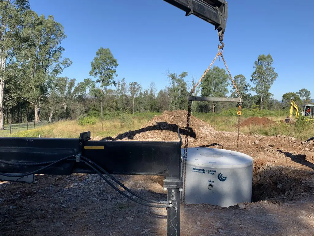 A crane is lifting a cylindrical concrete septic tank at a construction site surrounded by greenery. Trees and a small mound of earth are visible in the background under a clear blue sky. Equipment and machinery are partially seen in the scene, indicating both commercial and domestic use.