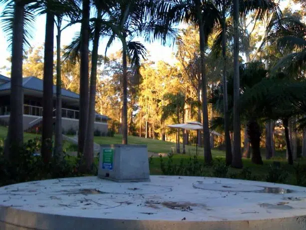 A circular concrete platform with debris is surrounded by tall trees and greenery. In the background, there is a building partially visible, and a patio with a table and chairs. The sky is clear, and the scene is bathed in warm sunlight, creating a serene domestic setting.